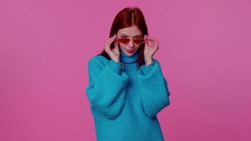 sincero frio alegre pelirrojo niña en azul suéter vistiendo Gafas de sol, encantador sonrisa en rosado pared video