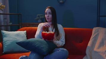 vrouw zittend Aan sofa alleen, aan het eten popcorn en aan het kijken interessant TV serieel, verschrikking film Bij huis video