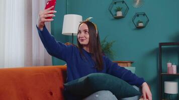 mujer en sofá a hogar con teléfono inteligente tomando selfie en móvil teléfono leva, virtual llamada en línea video