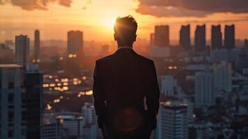lleno cuerpo retrato de exitoso empresario vistiendo un traje en pie cerca panorámico ventanas, mirando a puesta de sol terminado ciudad con rascacielos foto