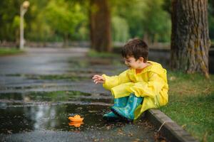 Child playing with toy boat in puddle. Kid play outdoor by rain. Fall rainy weather outdoors activity for young children. Kid jumping in muddy puddles. Waterproof jacket and boots for baby. childhood photo