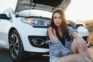 A young girl sits near a broken car on the road with an open hood. photo