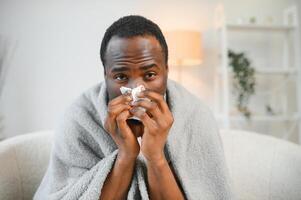 Sick mature black man covered in blanket sneezing runny nose while sitting on couch in living room, with lots of pills on table photo