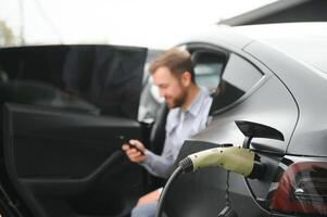 Happy man using smart phone and charging car at vehicle charging station photo