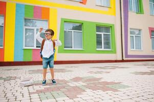espalda a escuela. contento sonriente chico en lentes es yendo a colegio para el primero tiempo. niño con mochila y libro al aire libre. comenzando de lecciones primero día de otoño foto