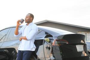 Stylish african man with coffe cup in hand inserts plug into the electric car charging socket photo