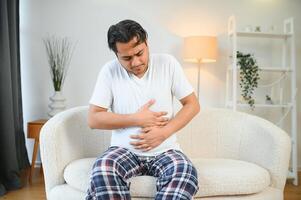 Sad exhausted guy of indian and arabian ethnicity, holds his hands on his stomach, grimaces from pain in his stomach, suffers from poisoning, spasm, stomach problems photo