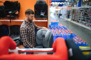 joven padre con elegir coche asiento en Tienda de para niños bienes foto