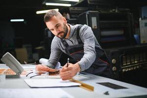 Printing house interior with modern offset print machine and operator in working uniform checking quality and controlling process of print photo