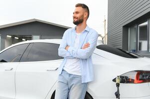 hermosa joven elegante hombre es con eléctrico coche a tiempo de día cargando el vehículo foto