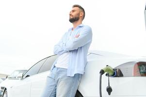 Beautiful young stylish man is with electric car at daytime charging the vehicle photo