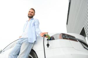 Beautiful young stylish man is with electric car at daytime charging the vehicle photo