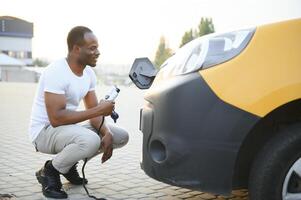 africano hombre participación cargar cable en en mano en pie cerca eléctrico coche. foto