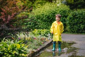 pequeño chico jugando en lluvioso verano parque. niño con paraguas, impermeable Saco y botas saltando en charco y barro en el lluvia. niño caminando en verano lluvia al aire libre divertido por ninguna clima. contento infancia. foto