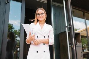 retrato de un exitoso mujer de negocios participación un digital tableta almohadilla y un carpeta mientras con orgullo en pie cerca oficina. foto