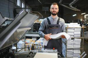 Printing house interior with modern offset print machine and operator in working uniform checking quality and controlling process of print photo