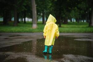 pequeño chico jugando en lluvioso verano parque. niño con paraguas, impermeable Saco y botas saltando en charco y barro en el lluvia. niño caminando en verano lluvia al aire libre divertido por ninguna clima. contento infancia. foto