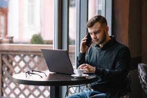 joven empresario hablando en móvil teléfono mientras trabajando en ordenador portátil en cafetería. foto