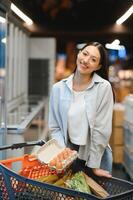 sonriente contento mujer disfrutando compras a el supermercado, ella es propensión en un lleno carro foto