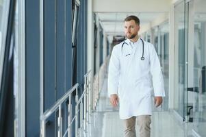 Portrait of handsome male doctor wearing white coat standing In hospital corridor. photo