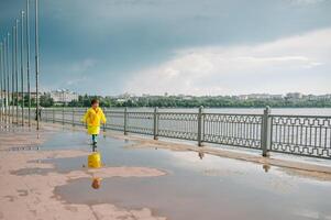 Little boy playing in rainy summer park. Child with umbrella, waterproof coat and boots jumping in puddle and mud in the rain. Kid walking in summer rain Outdoor fun by any weather. happy childhood. photo