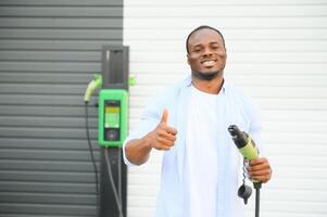 Serious african man holding charge cable in on hand, standing near luxury electric car. photo