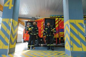 retrato de dos joven bomberos en uniforme en pie dentro el fuego estación foto