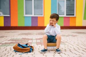 Sad little boy outside of school. Sad schoolboy with books near a modern school. School concept. Back to school. photo