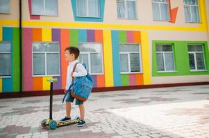 Adolescente chico con patada scooter cerca moderno escuela. niño con mochila y libro al aire libre. comenzando de lecciones primero día de caer. espalda a escuela. foto