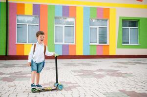 Adolescente chico con patada scooter cerca moderno escuela. niño con mochila y libro al aire libre. comenzando de lecciones primero día de caer. espalda a escuela. foto