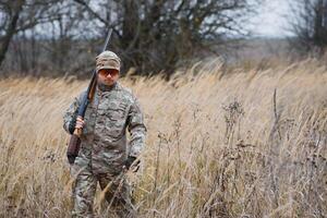 A male hunter with a gun while sitting takes aim at a forest. The concept of a successful hunt, an experienced hunter. Hunting the autumn season. The hunter has a rifle and a hunting uniform photo