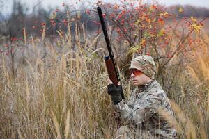 A male hunter with a gun while sitting takes aim at a forest. The concept of a successful hunt, an experienced hunter. Hunting the autumn season. The hunter has a rifle and a hunting uniform. photo