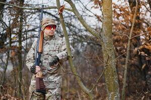 A male hunter with a gun while sitting takes aim at a forest. The concept of a successful hunt, an experienced hunter. Hunting the autumn season. The hunter has a rifle and a hunting uniform photo