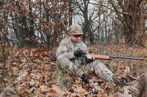 A male hunter with a gun while sitting takes aim at a forest. The concept of a successful hunt, an experienced hunter. Hunting the autumn season. The hunter has a rifle and a hunting uniform photo