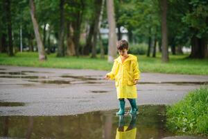 pequeño chico jugando en lluvioso verano parque. niño con paraguas, impermeable Saco y botas saltando en charco y barro en el lluvia. niño caminando en verano lluvia al aire libre divertido por ninguna clima. contento infancia. foto