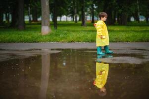 pequeño chico jugando en lluvioso verano parque. niño con paraguas, impermeable Saco y botas saltando en charco y barro en el lluvia. niño caminando en verano lluvia al aire libre divertido por ninguna clima. contento infancia. foto