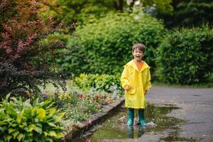 Little boy playing in rainy summer park. Child with umbrella, waterproof coat and boots jumping in puddle and mud in the rain. Kid walking in summer rain Outdoor fun by any weather. happy childhood. photo