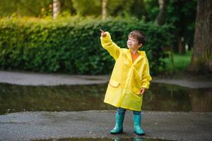 Little boy playing in rainy summer park. Child with umbrella, waterproof coat and boots jumping in puddle and mud in the rain. Kid walking in summer rain Outdoor fun by any weather. happy childhood photo