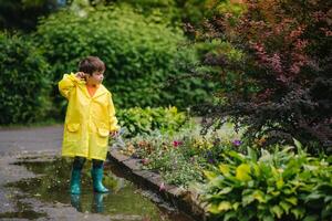Little boy playing in rainy summer park. Child with umbrella, waterproof coat and boots jumping in puddle and mud in the rain. Kid walking in summer rain Outdoor fun by any weather. happy childhood. photo