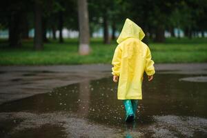 Little boy playing in rainy summer park. Child with umbrella, waterproof coat and boots jumping in puddle and mud in the rain. Kid walking in summer rain Outdoor fun by any weather. happy childhood. photo