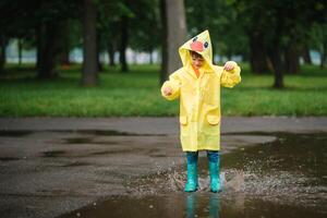 pequeño chico jugando en lluvioso verano parque. niño con paraguas, impermeable Saco y botas saltando en charco y barro en el lluvia. niño caminando en verano lluvia al aire libre divertido por ninguna clima. contento infancia. foto