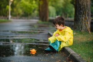 Child playing with toy boat in puddle. Kid play outdoor by rain. Fall rainy weather outdoors activity for young children. Kid jumping in muddy puddles. Waterproof jacket and boots for baby. childhood photo