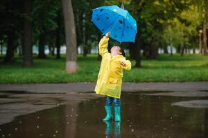 Child playing with toy boat in puddle. Kid play outdoor by rain. Fall rainy weather outdoors activity for young children. Kid jumping in muddy puddles. Waterproof jacket and boots for baby. childhood photo