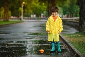 Child playing with toy boat in puddle. Kid play outdoor by rain. Fall rainy weather outdoors activity for young children. Kid jumping in muddy puddles. Waterproof jacket and boots for baby. childhood photo