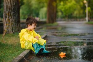 niño jugando con juguete barco en charco. niño jugar al aire libre por lluvia. otoño lluvioso clima al aire libre actividad para joven niños. niño saltando en lodoso charcos impermeable chaqueta y botas para bebé. infancia foto