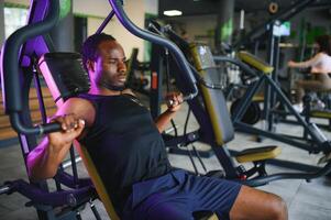 atlético joven negro hombre teniendo rutina de ejercicio en cofre prensa máquina en gimnasio foto