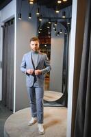 moderno joven hermoso empresario vestido en clásico traje ajustando un Corbata en frente de el espejo mientras en pie en el traje tienda foto