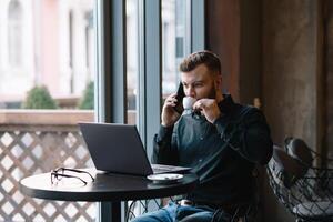 Young businessman talking on mobile phone while working on laptop in cafe photo