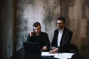 dos empresarios en un oficina sonriente a el cámara mientras trabajando juntos detrás un ordenador portátil computadora. foto