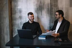 dos empresarios en un oficina sonriente a el cámara mientras trabajando juntos detrás un ordenador portátil computadora. foto